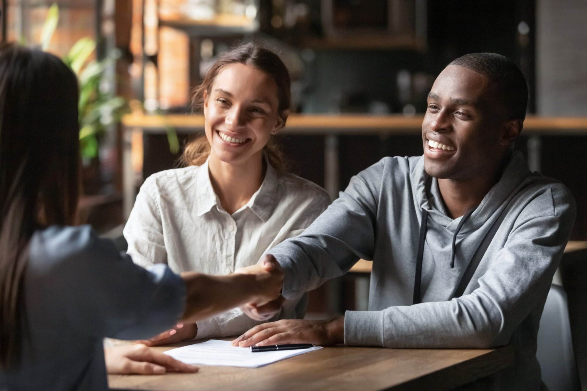 Diverse couple smiling and shaking hands with a professional across a table in a cozy office setting.