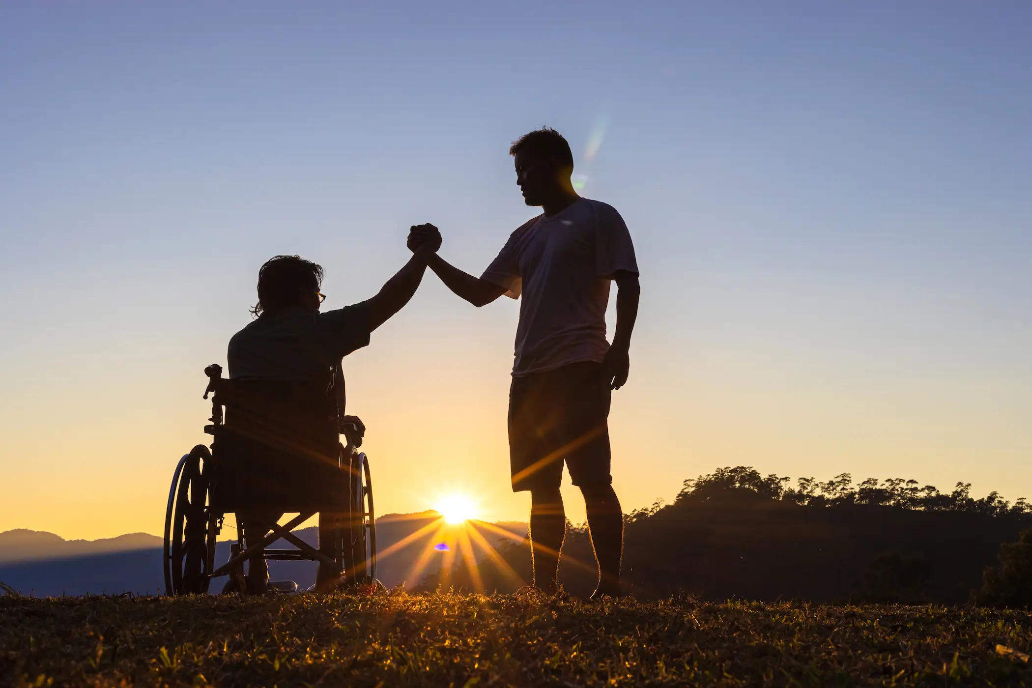 Silhouette of a person in a wheelchair and another standing, sharing a moment at sunrise.