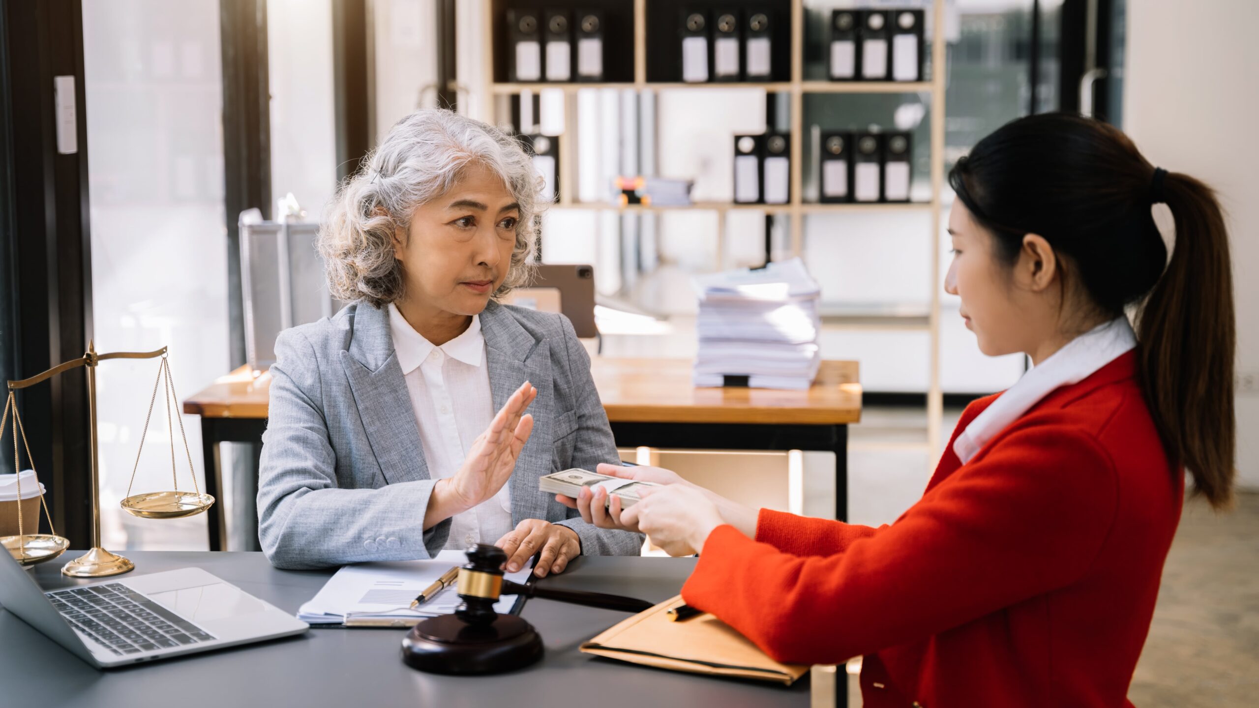 Two women, one elderly and one younger, in a legal consultation setting. The elderly woman is refusing a stack of money offered by the younger woman.