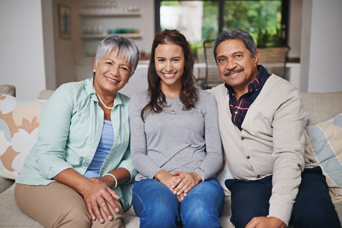 A happy family sitting together on a couch, featuring a smiling woman with her older parents in a cozy living room.